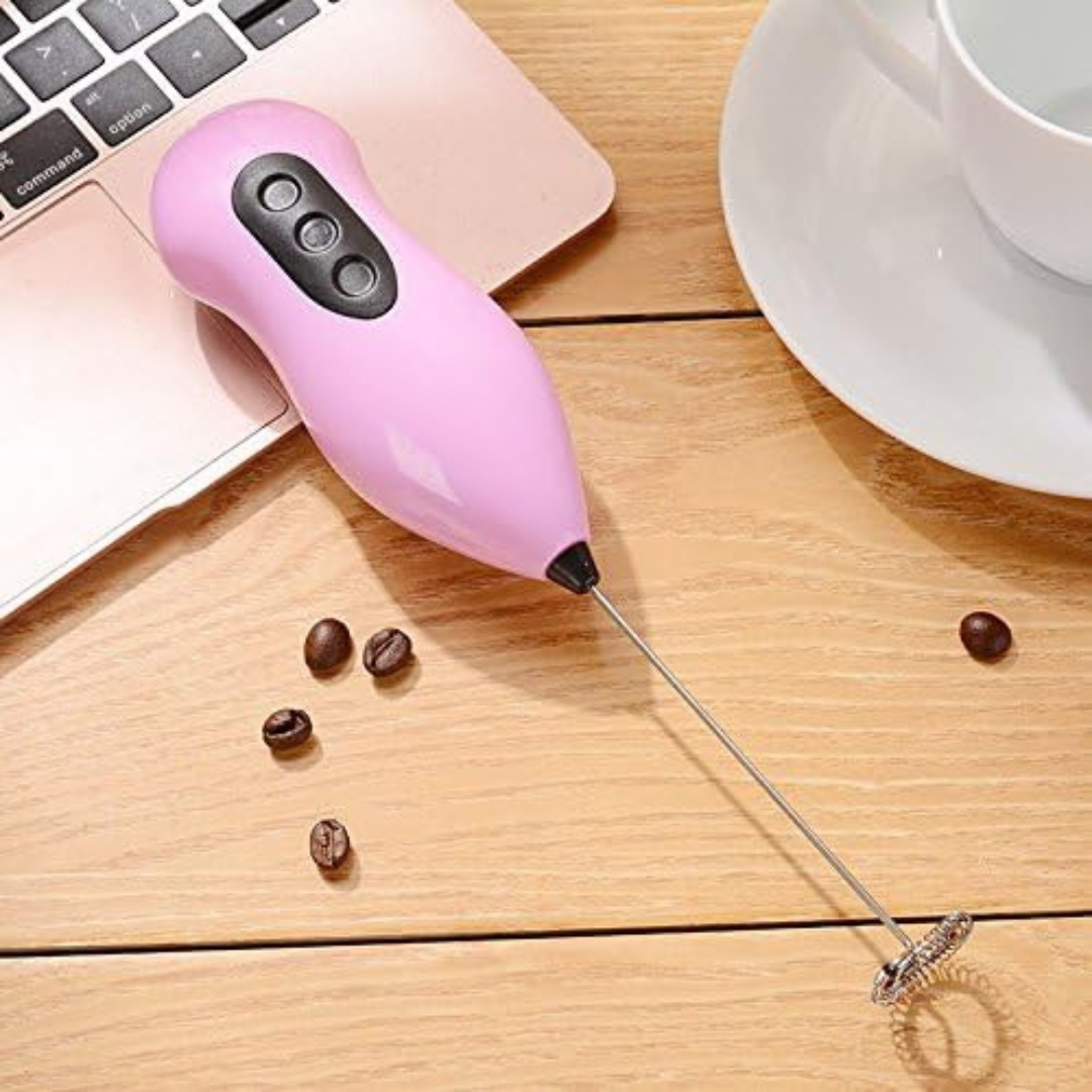 Pink handheld milk frother on a wooden surface with coffee beans and a laptop.