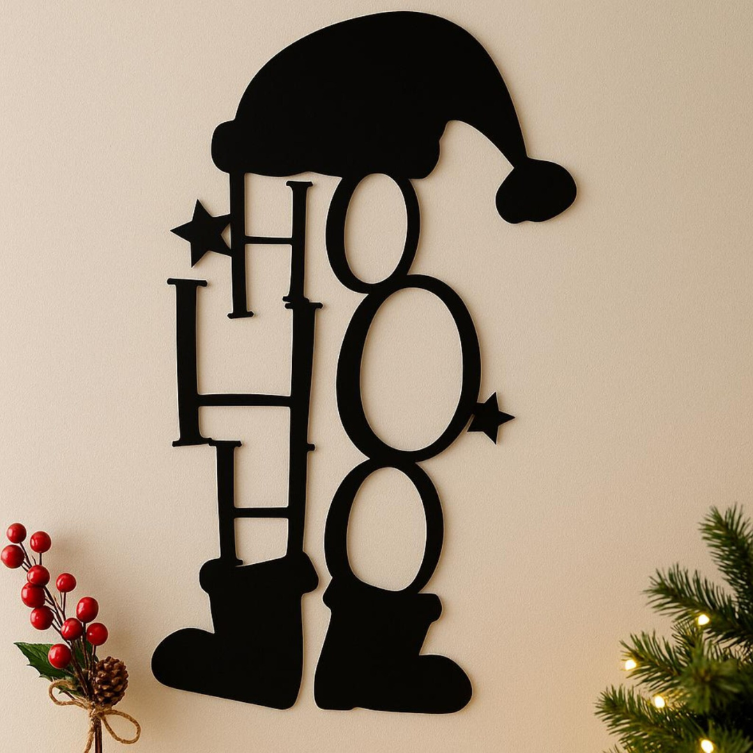 Black 'HO HO HO' wall decoration with Santa hat on a beige wall, Christmas tree and holly in the corner.
